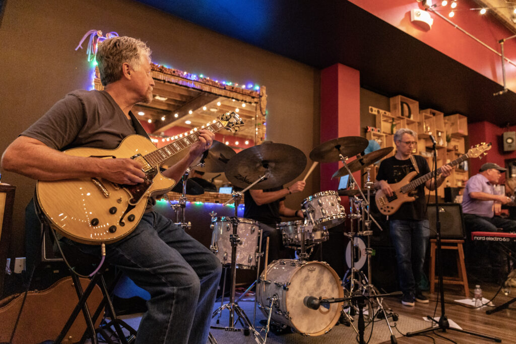 3 white men on a small stage. Two of them are playing electric guitars on the sides and in the middle there is a man playin on the drums. The environment is dimly lit with colorful string lights behind the.