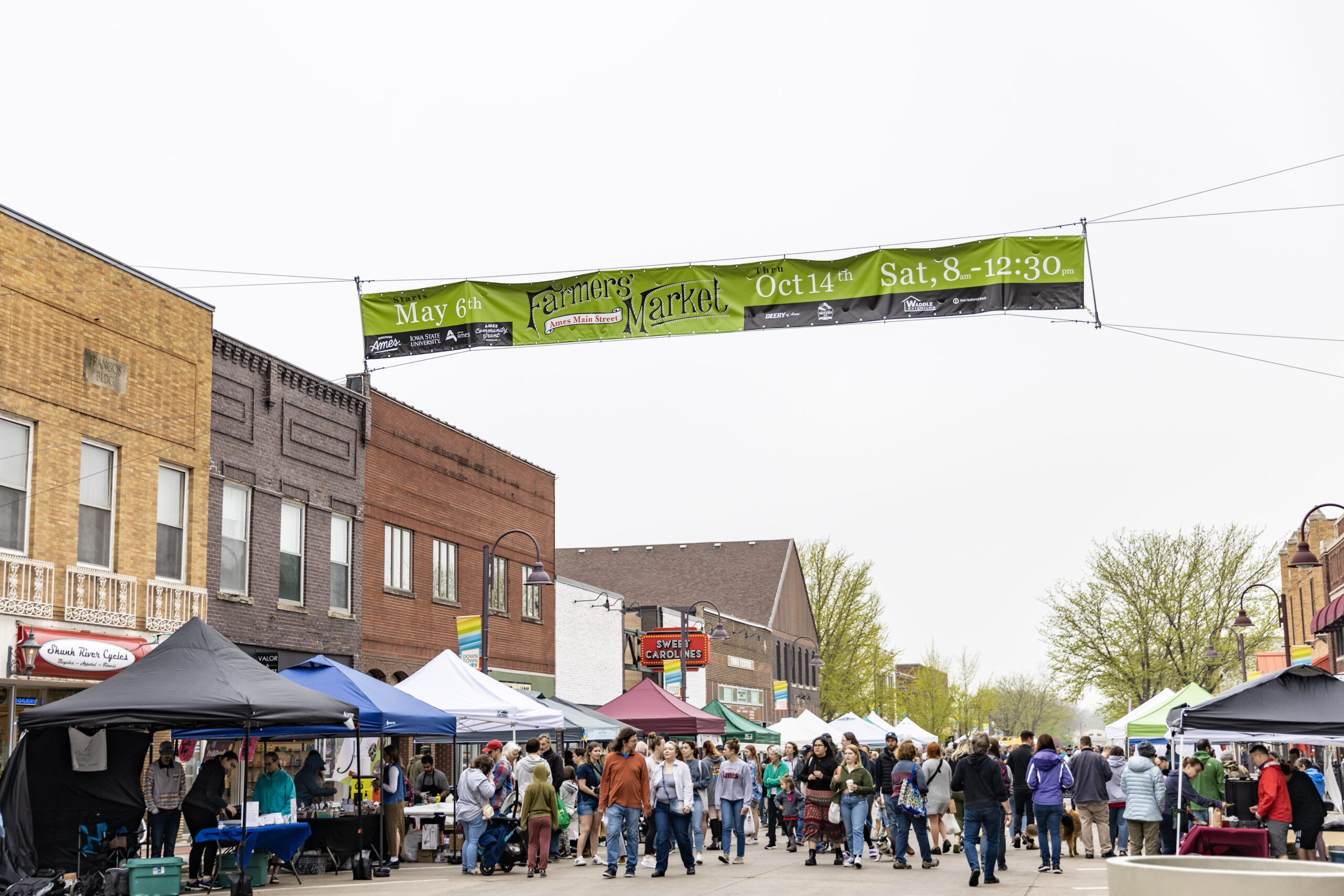 Ames Main Street Farmers Market from afar with people and tens and a big sign.