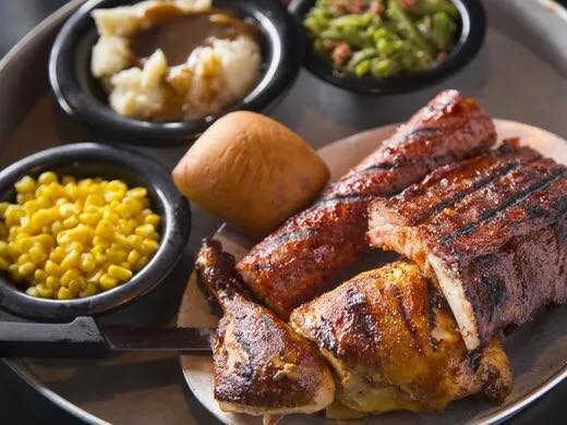 Hickory Park barbeque meal. Plate with sausage, pork tenderloin, ribs, and chicken. A bowl of corn, bowl of mashed potato and gravy and a bowl of green beans and bacon bits. There is a roll on the plate of meat. 