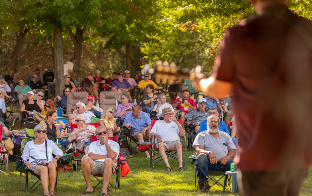 Man facing away from camera towards a crowd. He is wearing a red shirt and playing the guitar. The photo is outdoors. The crowd are sitting in lawn chairs in the grass and there are trees behind them.