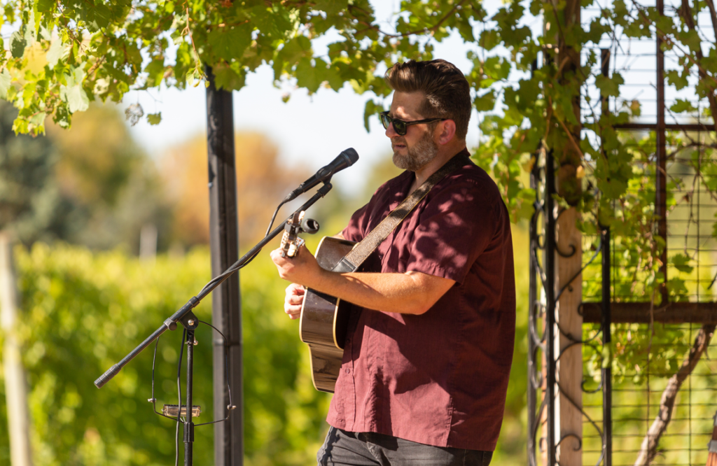 Guy in red shirt with guitar performing and singing out outside.