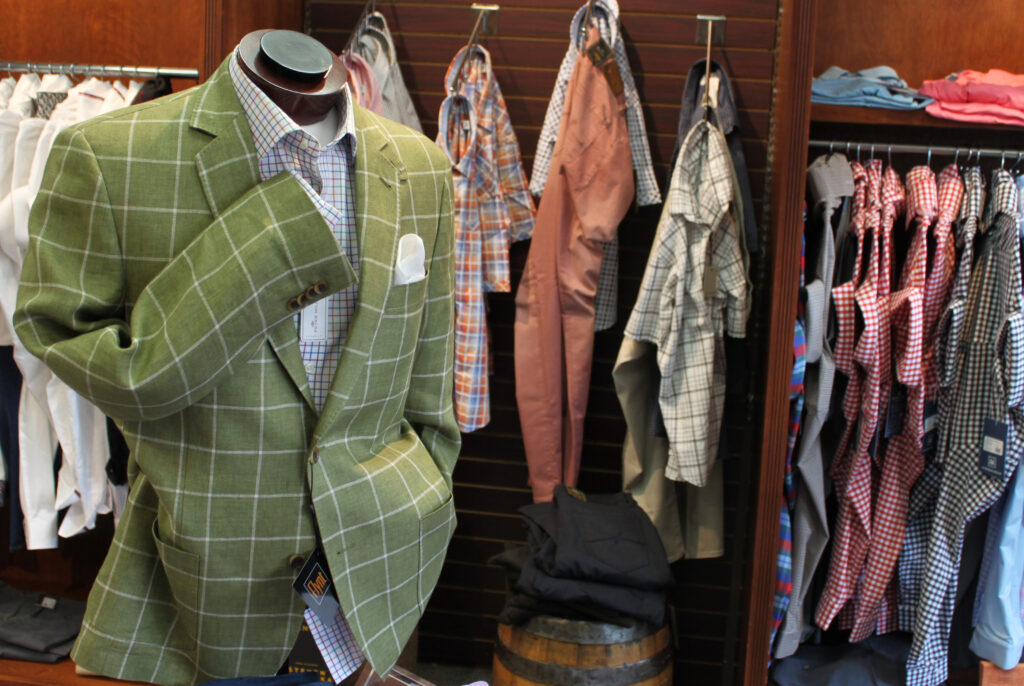 Green suit on mannequin. Light blue undershirt. Behind the mannequin there are more button downs and dress shirts on a brown wall. 