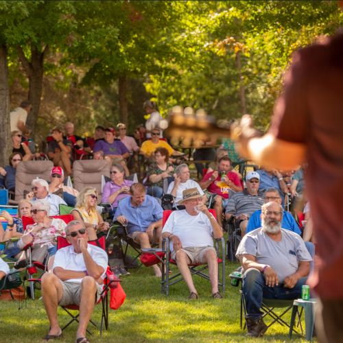 Man facing away from camera towards a crowd. He is wearing a red shirt and playing the guitar. The photo is outdoors. The crowd are sitting in lawn chairs in the grass and there are trees behind them.