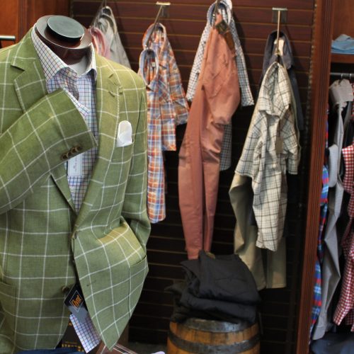 Green suit on mannequin. Light blue undershirt. Behind the mannequin there are more button downs and dress shirts on a brown wall.